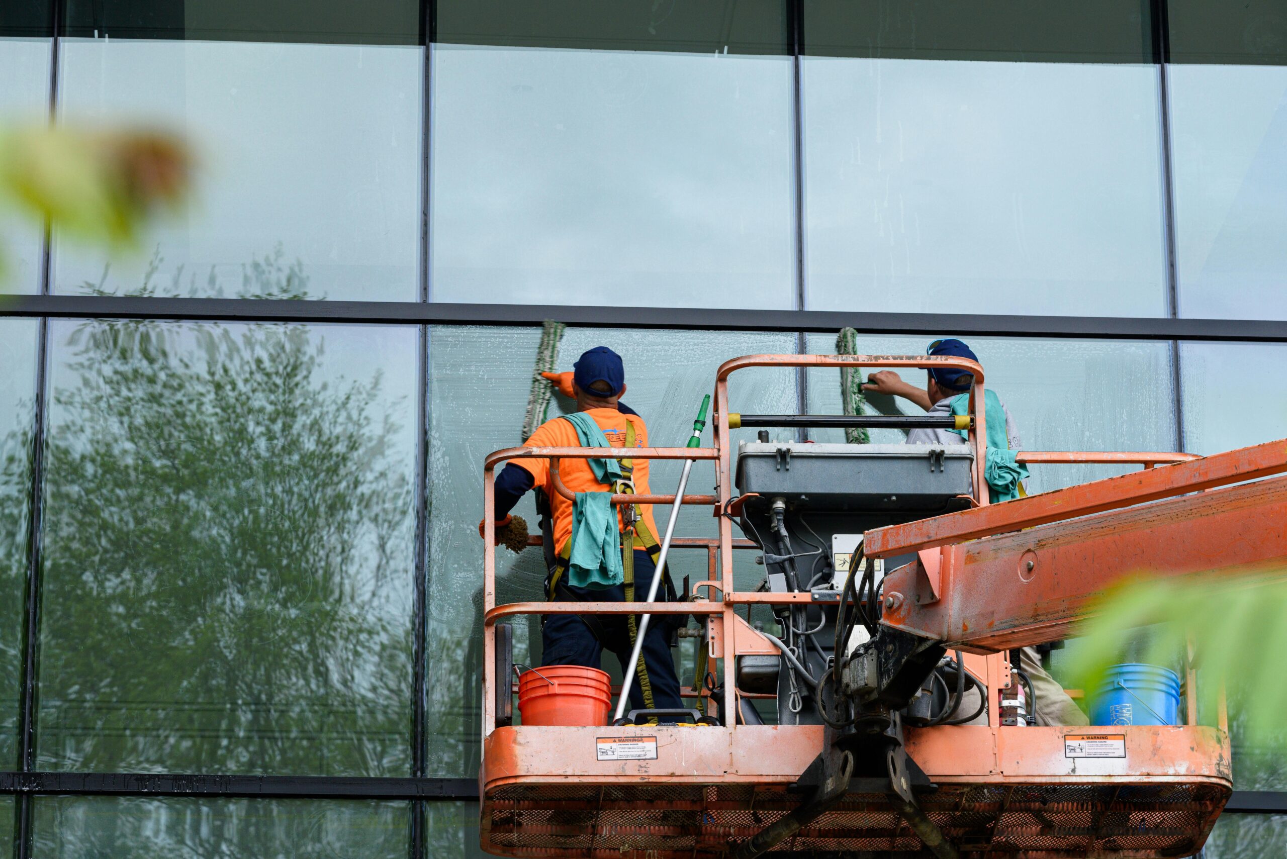 pexels-photo-31983866-31983866 Two workers cleaning windows on a glass building facade using a lift platform, enhancing building aesthetics.