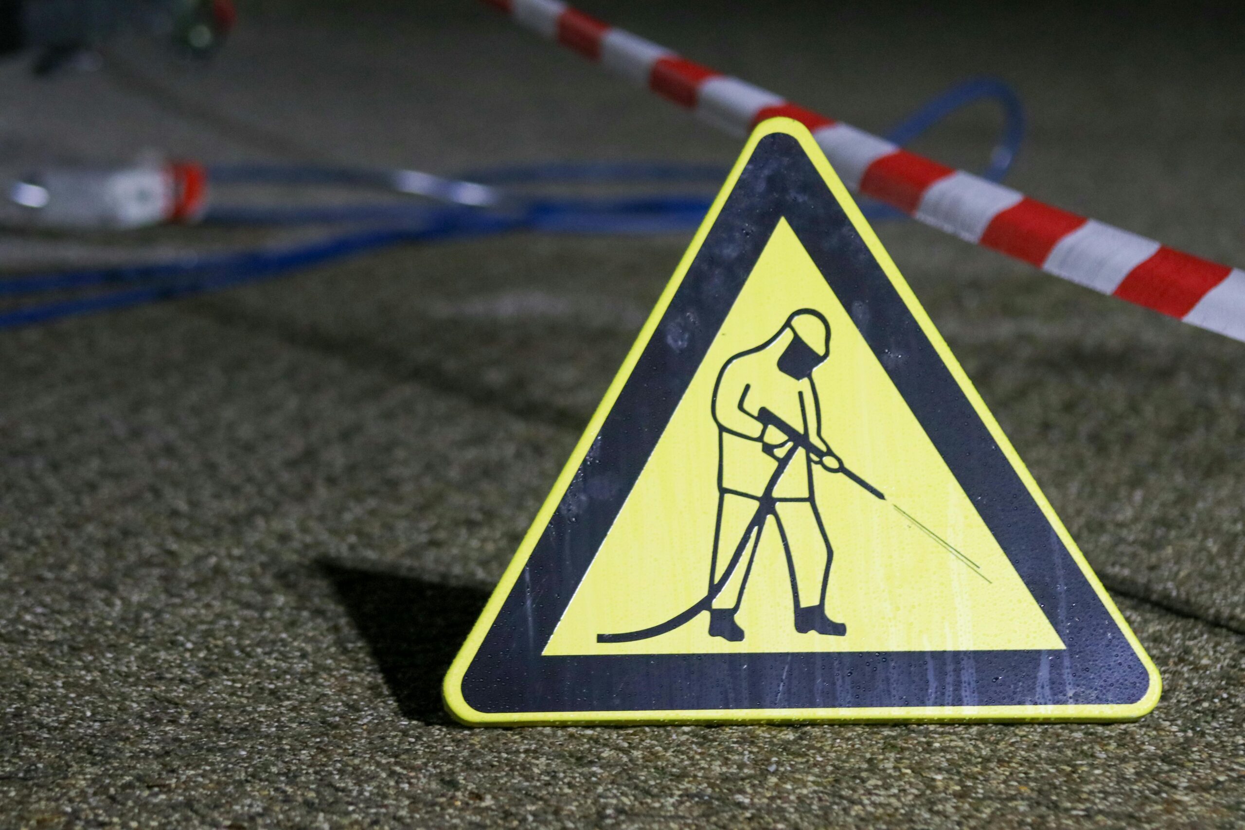 Yellow caution sign depicting pressure washing in a night setting with safety barriers.
